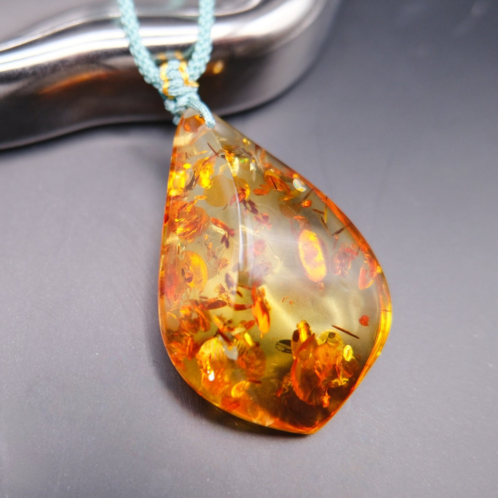 Pendant: golden Baltic amber teardrop with orange botanical inclusions, teal cord, by a metal tray