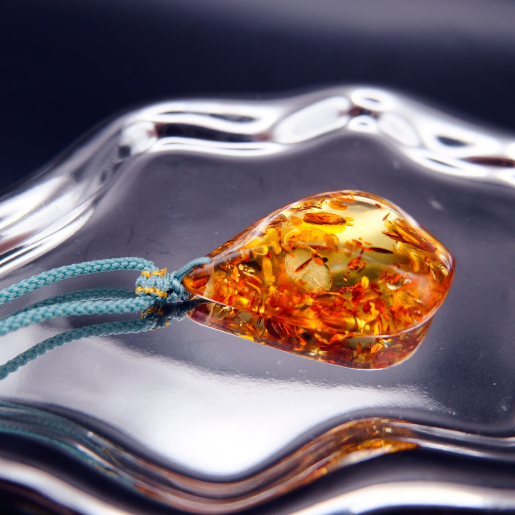 Pendant: golden Baltic amber teardrop with orange botanical inclusions, teal cord, lying flat on a mirrored surface