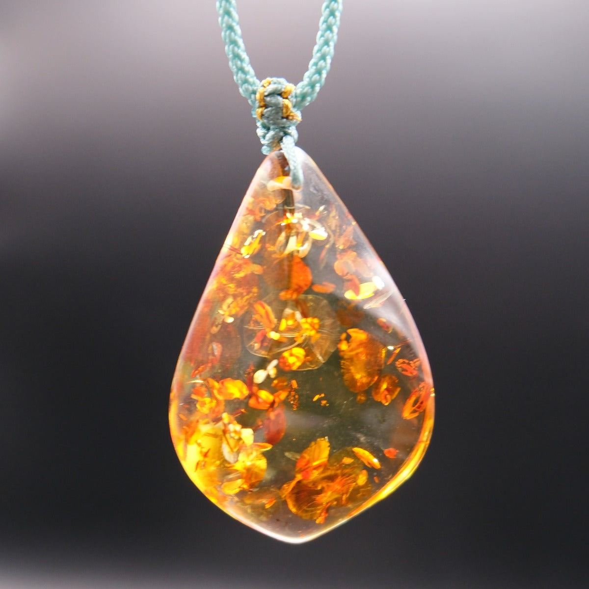Pendant: golden Baltic amber teardrop with orange botanical inclusions, teal cord, hanging against a dark background