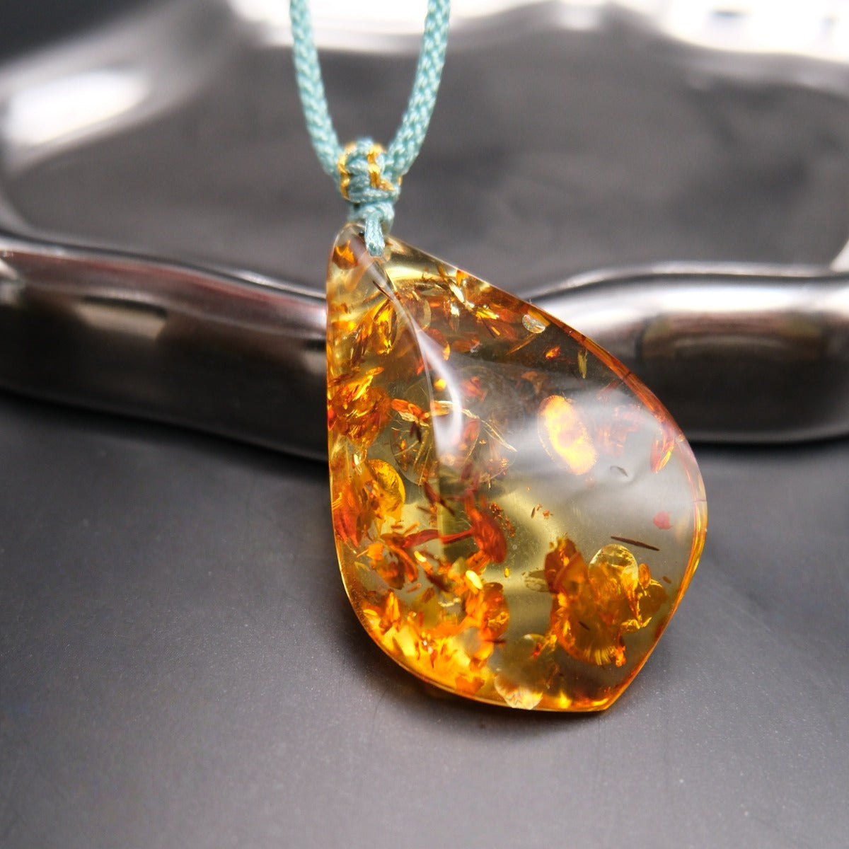 Pendant: golden Baltic amber teardrop with orange botanical inclusions, teal cord, leaning on a metal tray