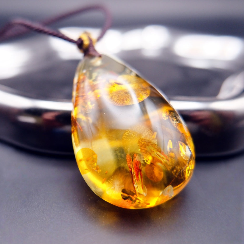 Pendant: golden Baltic amber teardrop with round botanical inclusions, dark cord, leaning on a metal tray