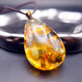 Pendant: golden Baltic amber teardrop with round botanical inclusions, dark cord, leaning on a metal tray