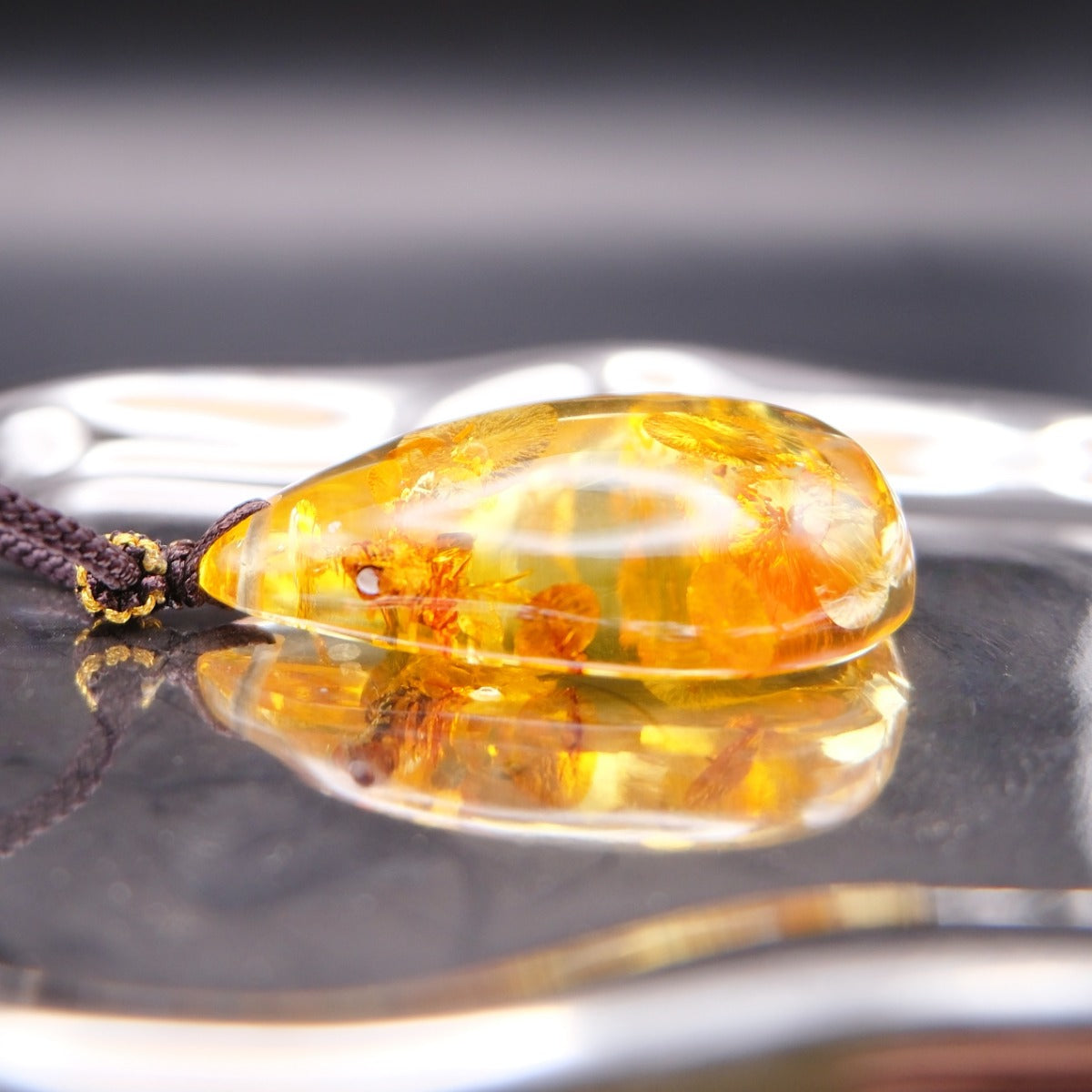 Pendant: golden Baltic amber teardrop with round botanical inclusions, dark cord, lying flat on a mirrored surface