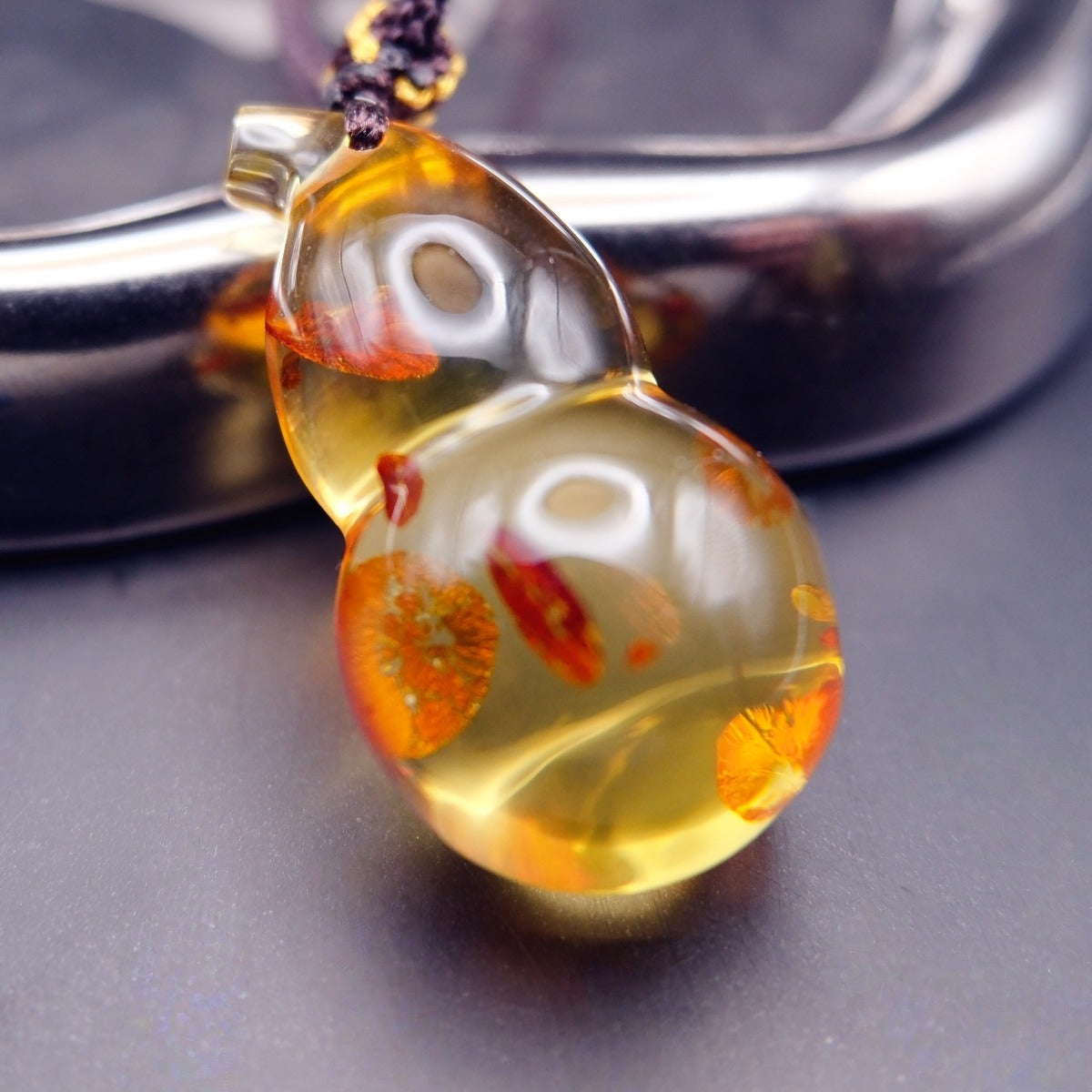 Pendant: golden Baltic amber gourd with orange-red inclusions, dark braided cord, leaning on a metal tray
