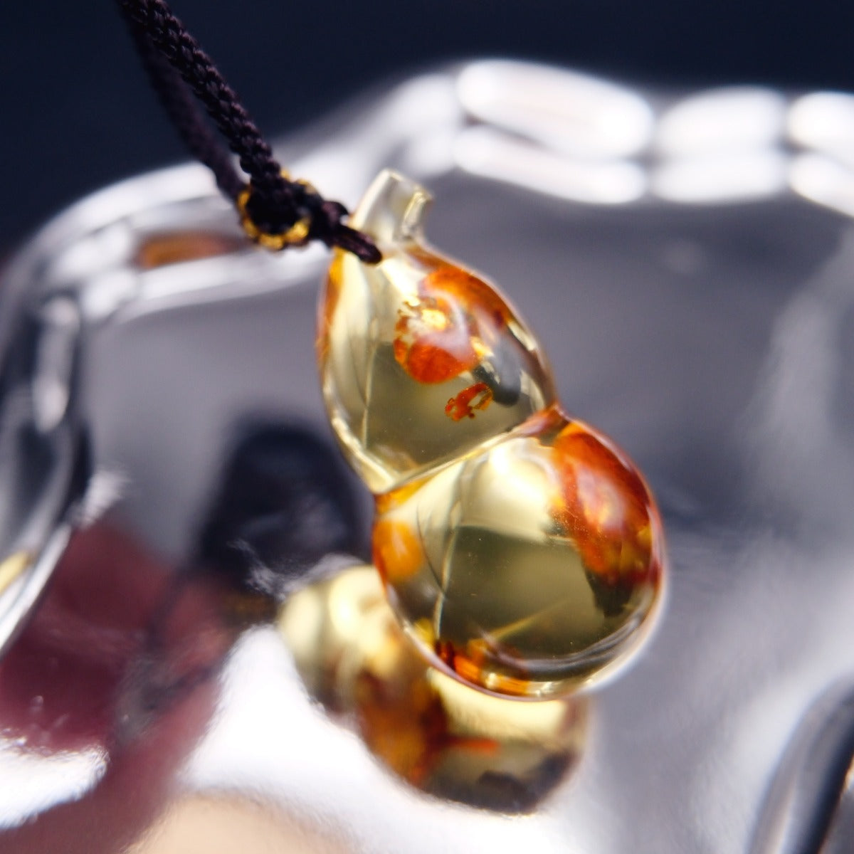 Pendant: golden Baltic amber gourd with orange-red inclusions, dark braided cord, side view on a mirrored surface