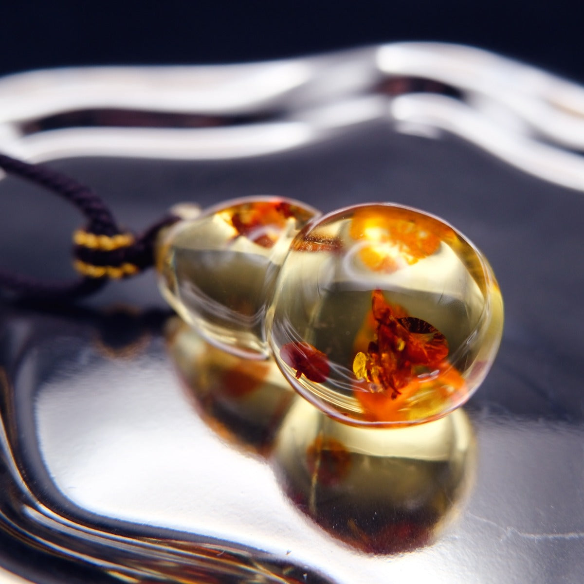 Pendant: golden Baltic amber gourd with orange-red inclusions, dark braided cord, lying on a mirrored surface