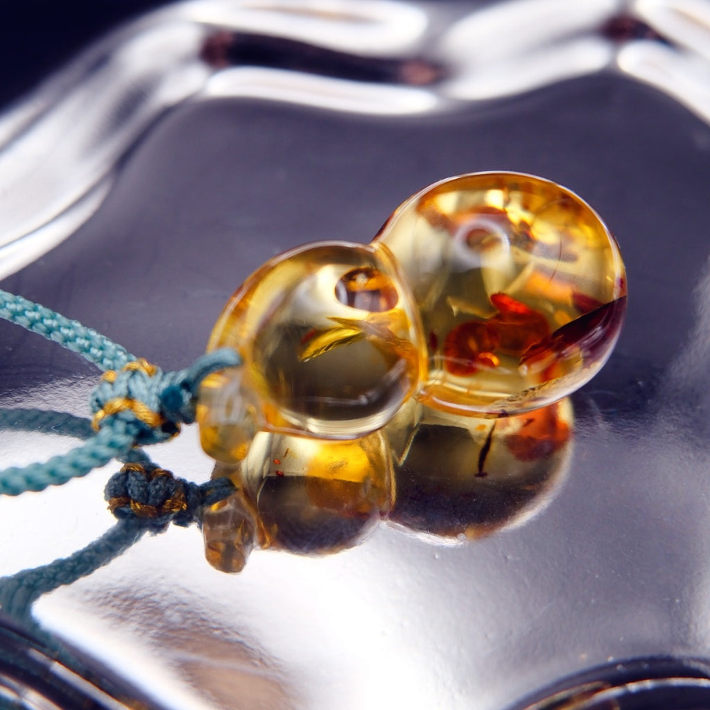 Pendant: transparent golden Baltic amber gourd with pale and orange inclusions, teal cord, lying flat on a mirrored surface