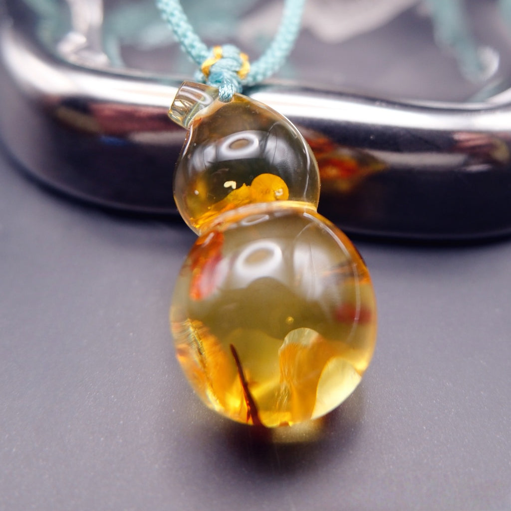 Pendant: transparent golden Baltic amber gourd with pale and orange inclusions, teal cord, standing against a metal tray