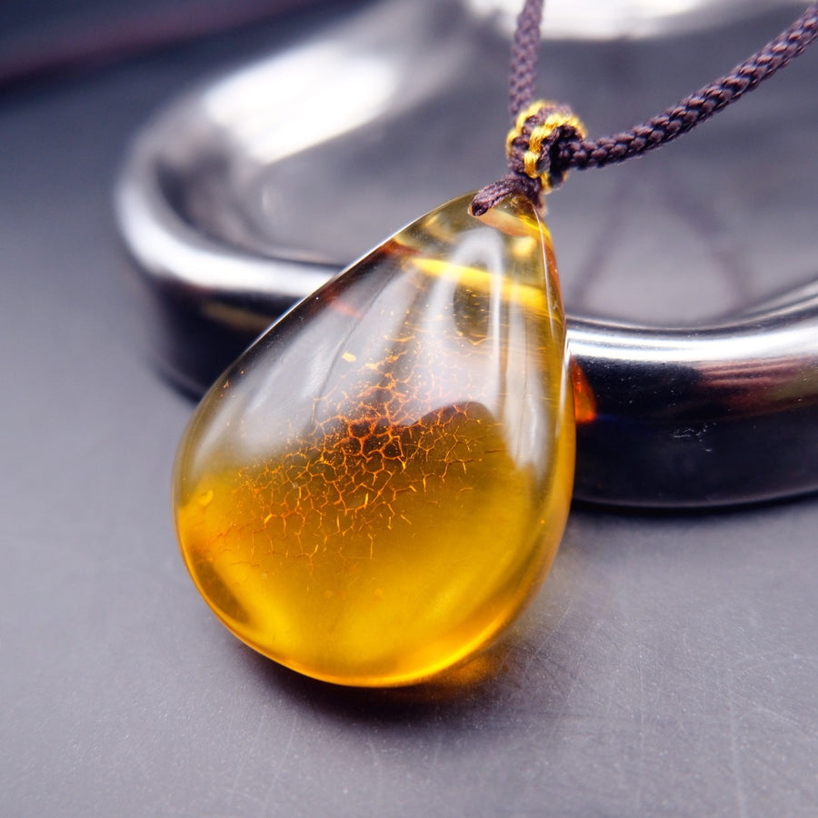 Pendant: transparent golden-yellow Baltic amber teardrop with a crackle-pattern inclusion, dark cord, leaning on a tray