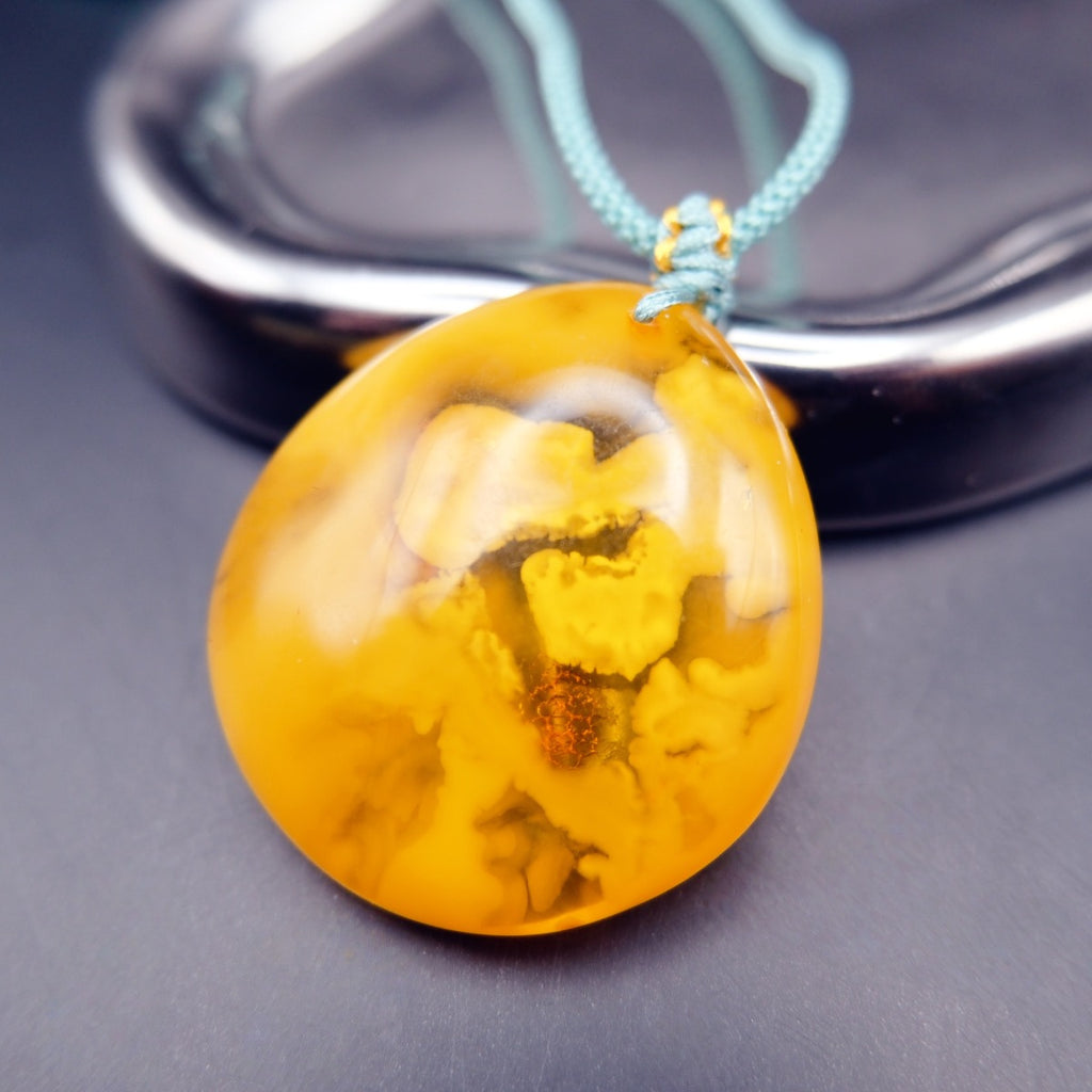 Pendant: opaque yellow-orange Baltic amber with paler cloud-like patches, teal cord, leaning on a metal tray