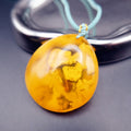 Pendant: opaque yellow-orange Baltic amber with paler cloud-like patches, teal cord, leaning on a metal tray