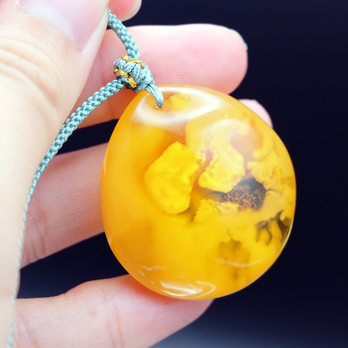 Pendant: opaque yellow-orange Baltic amber with paler cloud-like patches, teal cord, held between fingers