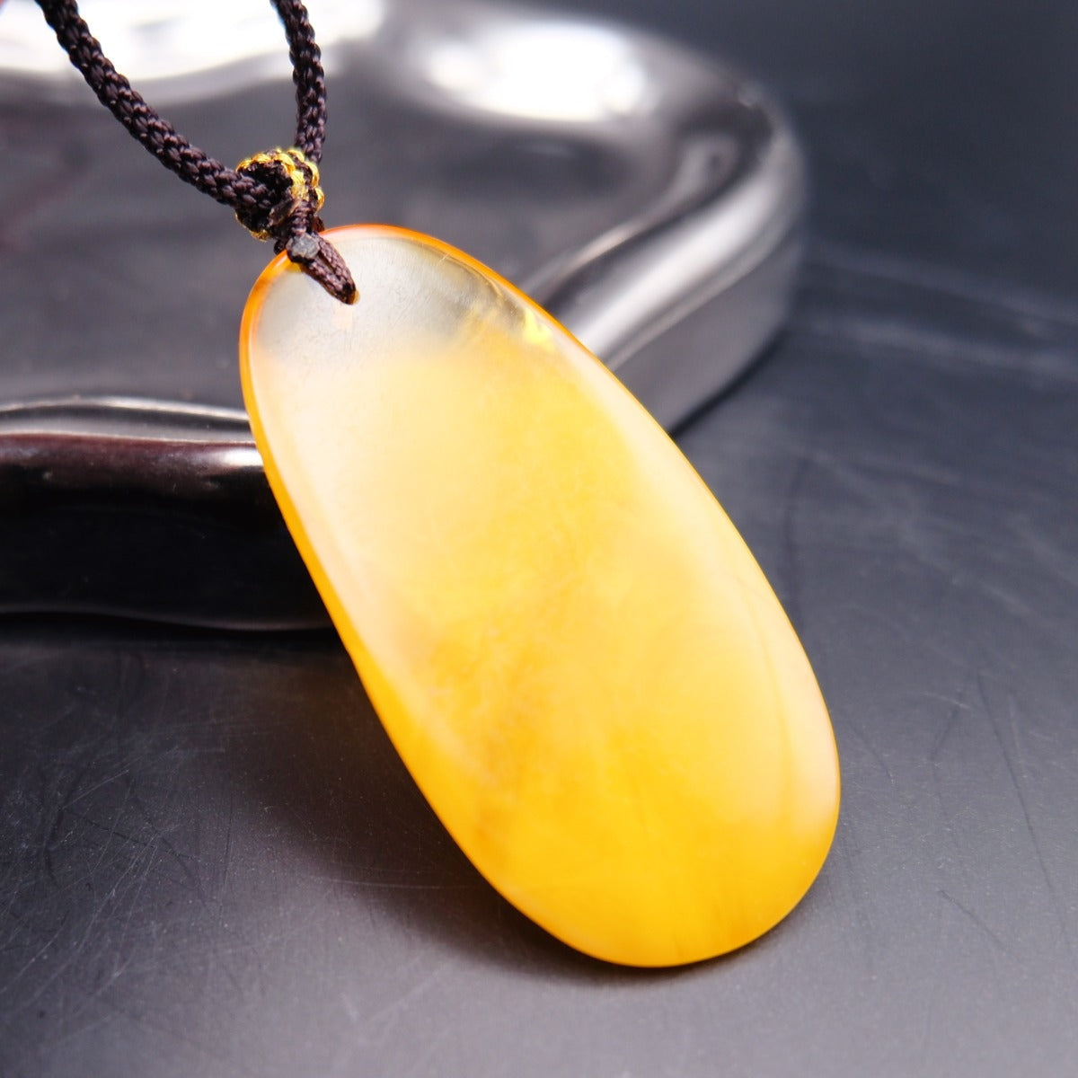 Pendant: long opaque yellow Baltic amber drop with a clear top edge, dark cord, leaning on a metal tray