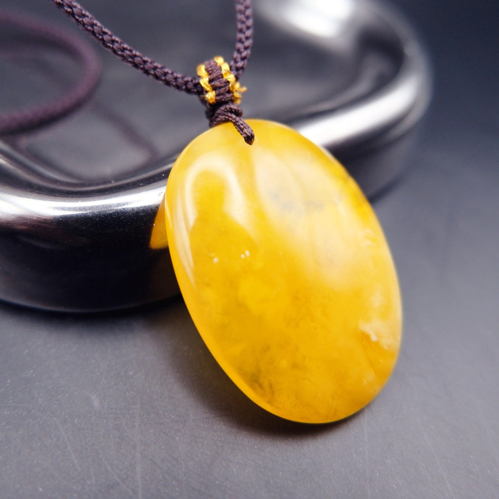 Pendant: yellow Baltic amber oval with cloudy white patches, dark cord, leaning on a metal tray