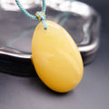 Pendant: pale opaque yellow Baltic amber oval drop, teal cord, leaning on a metal tray