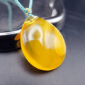 Pendant: yellow Baltic amber teardrop, translucent top and opaque lower half, teal cord, leaning on a metal tray