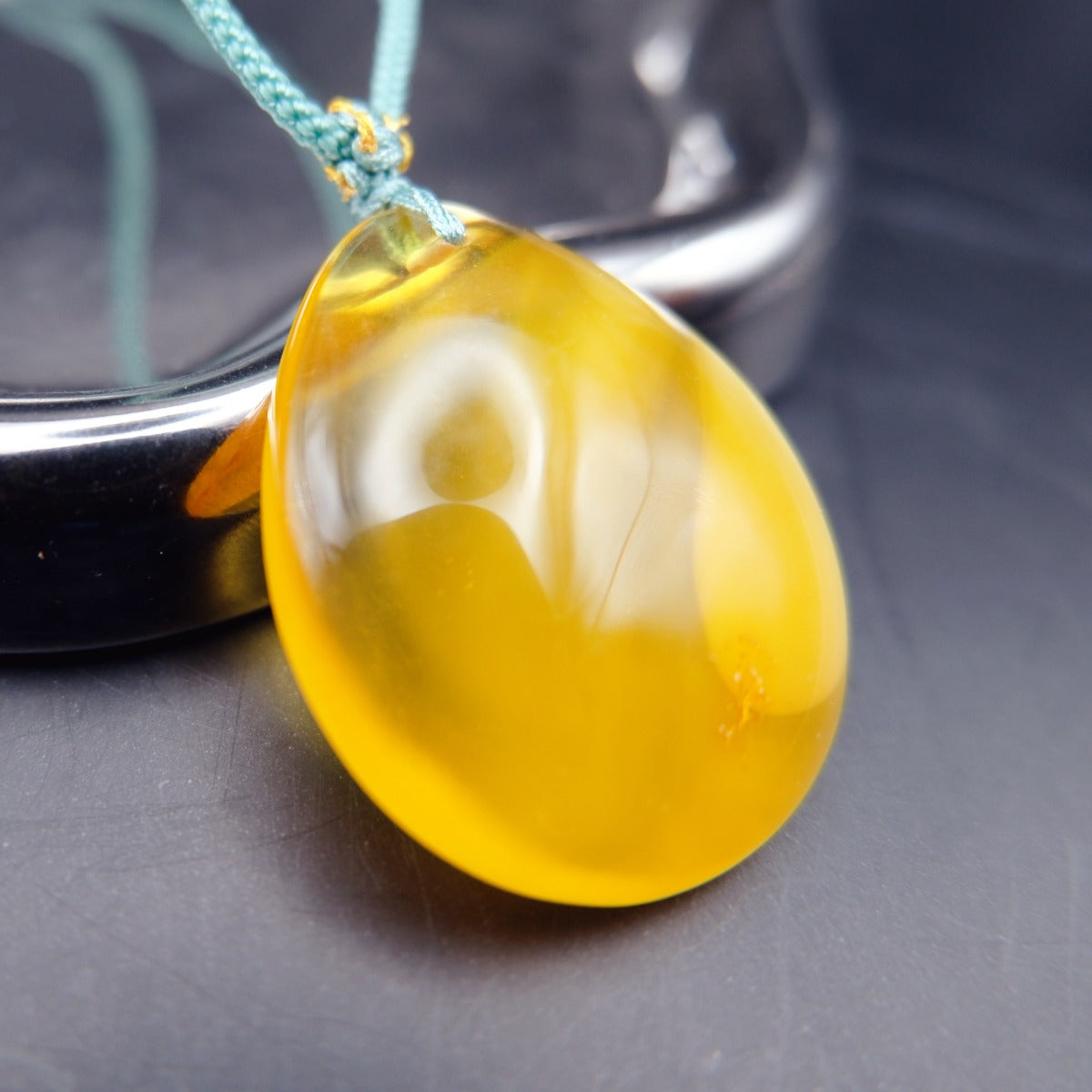 Pendant: yellow Baltic amber teardrop, translucent top and opaque lower half, teal cord, leaning on a metal tray