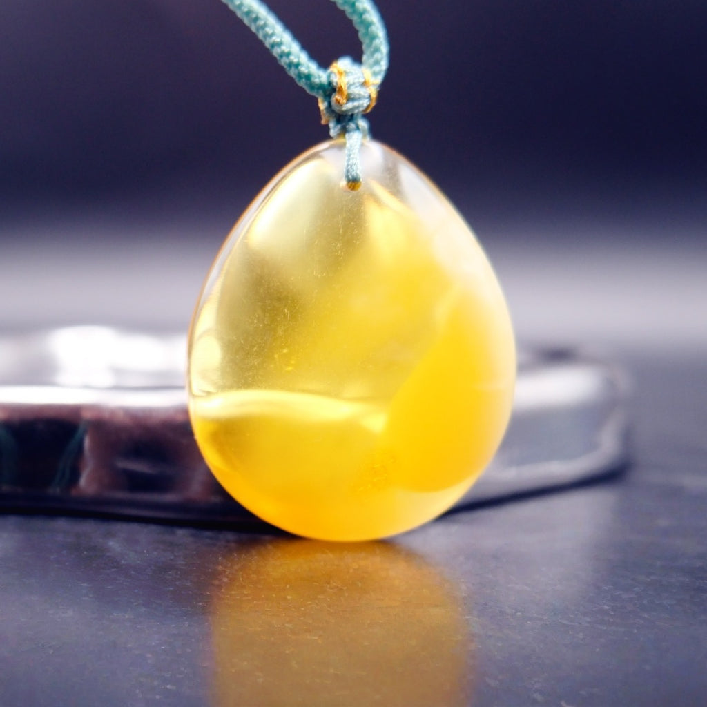 Pendant: yellow Baltic amber teardrop, translucent top and opaque lower half, teal cord, standing on a dark surface