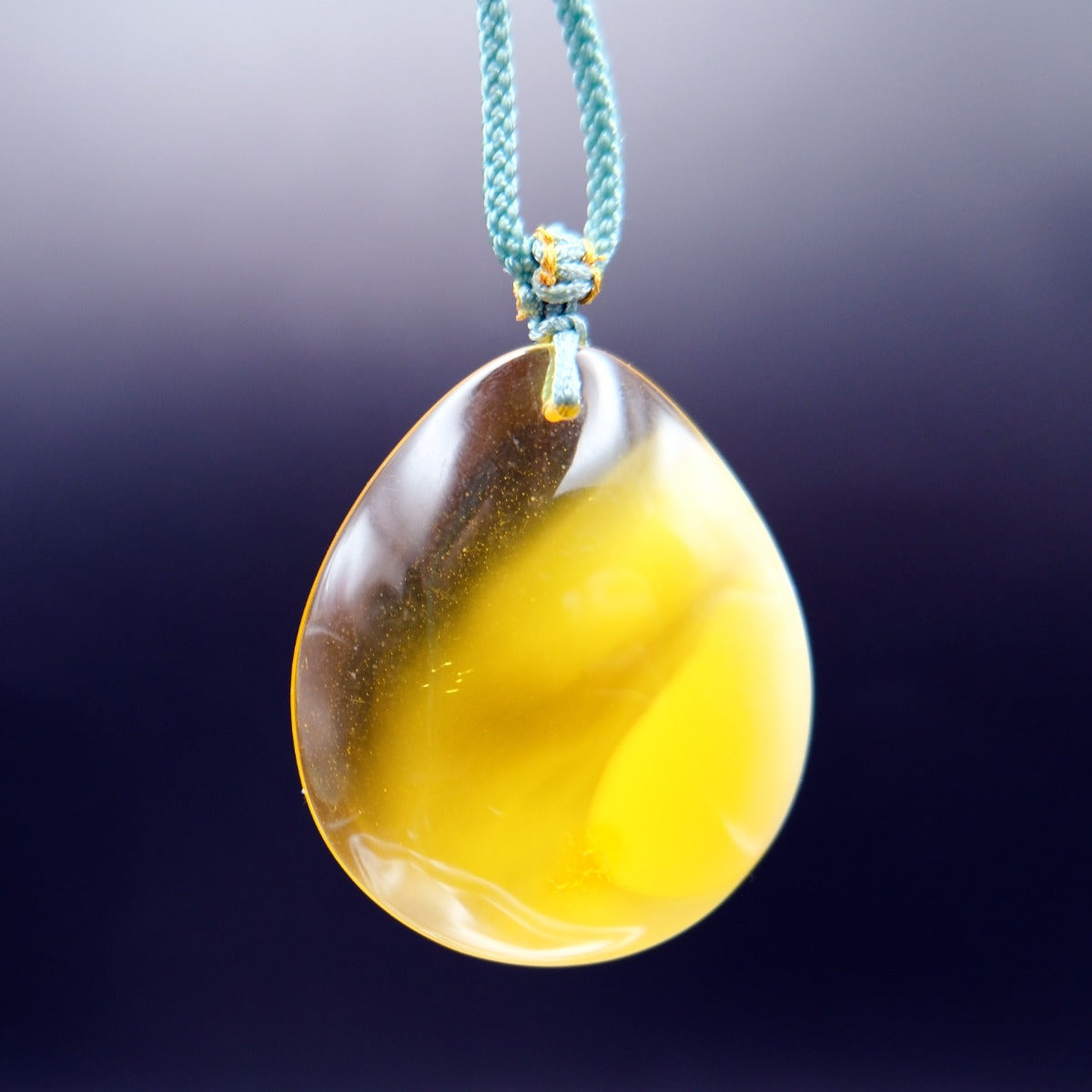 Pendant: yellow Baltic amber teardrop, translucent top and opaque lower half, teal cord, hanging against a dark background