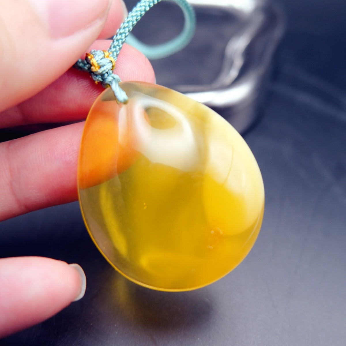 Pendant: yellow Baltic amber teardrop, translucent top and opaque lower half, teal cord, held in fingers