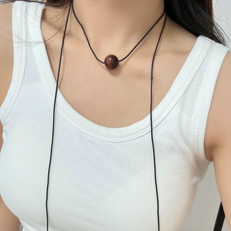 Pendant necklace worn as a choker: brown-red tiger's eye bead on a black cord, the long cord ends hanging over a white top