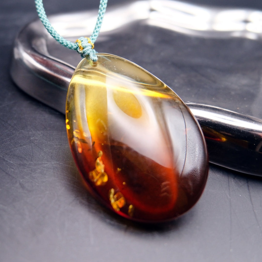 Pendant: transparent Baltic amber teardrop, yellow shading to dark red, golden flakes, teal cord, leaning on a tray