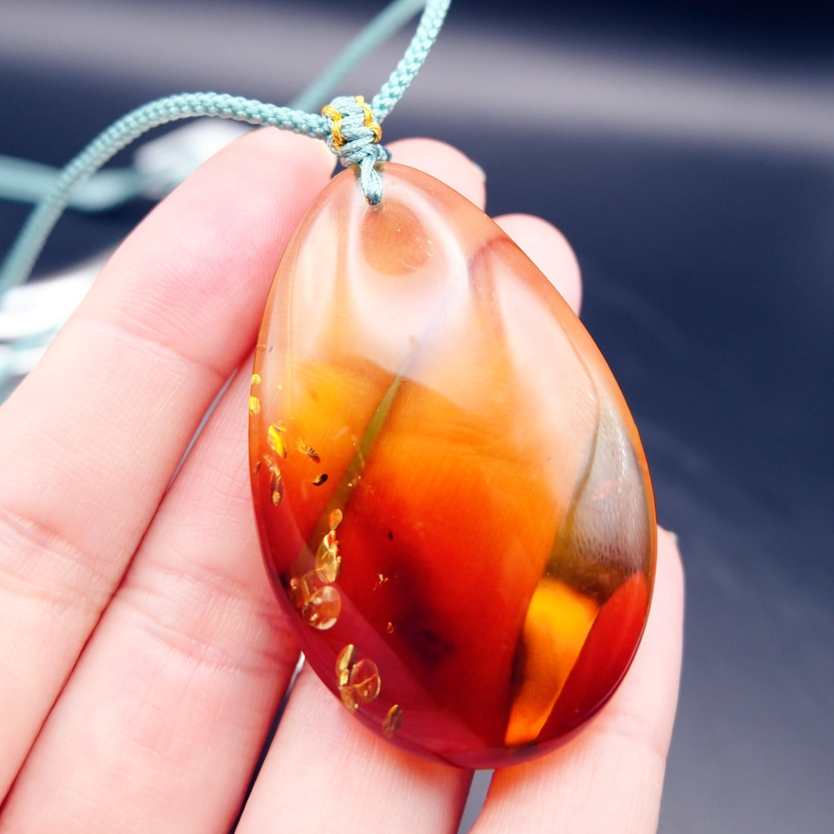 Pendant: transparent Baltic amber teardrop, yellow shading to dark red, golden flakes, teal cord, held on fingers