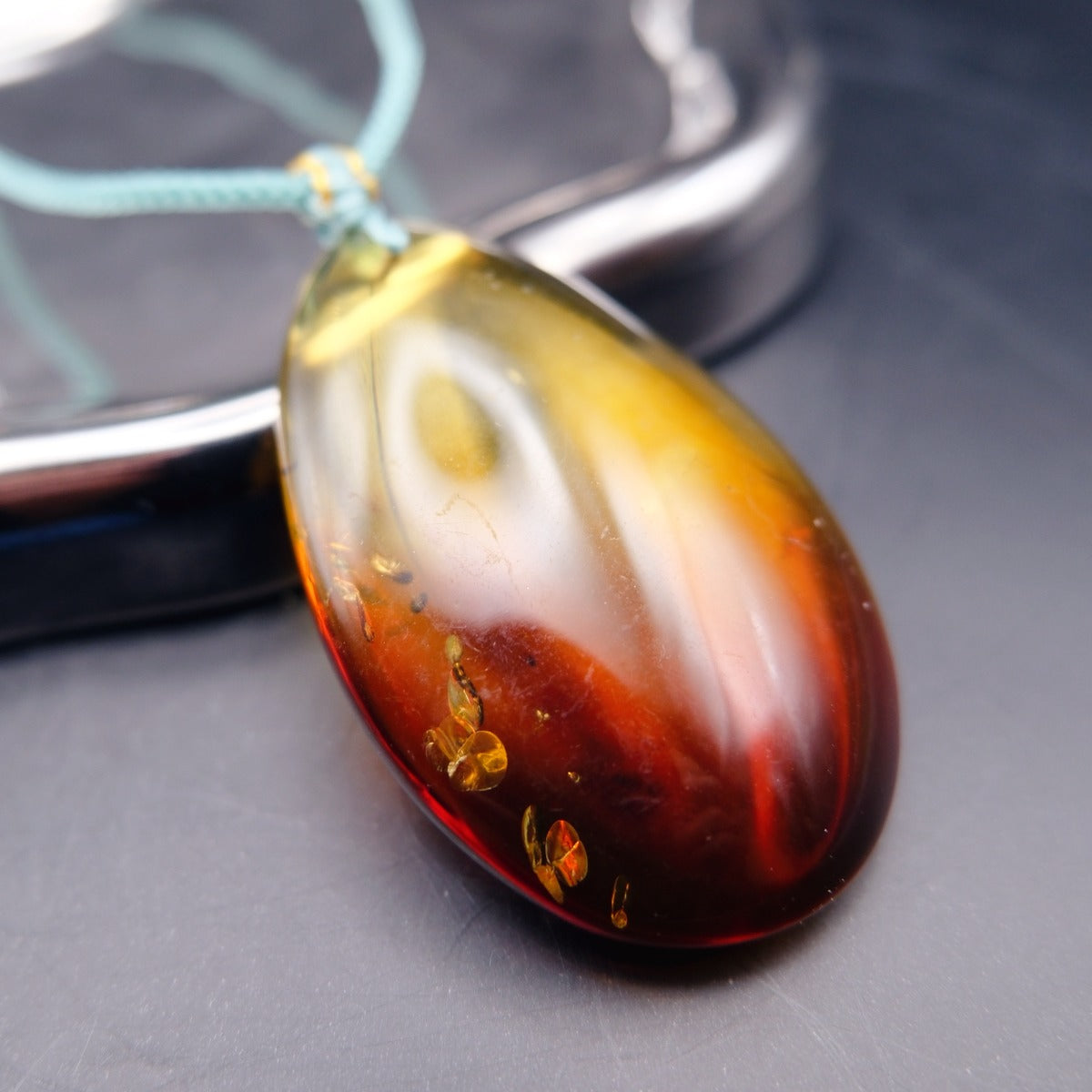 Pendant: transparent Baltic amber teardrop, yellow shading to dark red, golden flakes, teal cord, lying on a dark surface