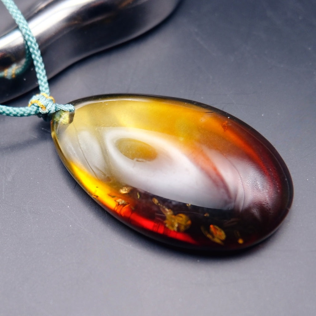 Pendant: transparent Baltic amber teardrop, yellow shading to dark red, golden flakes, teal cord, lying, side view