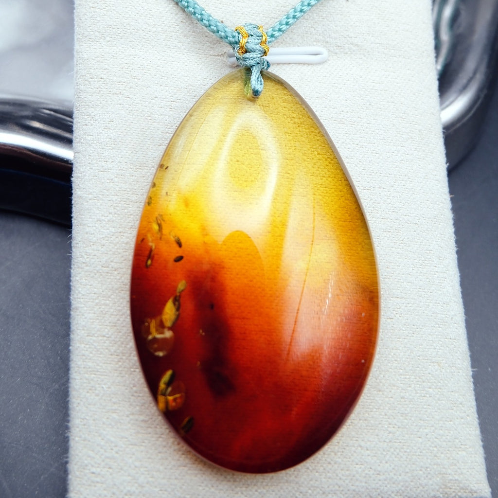 Pendant: transparent Baltic amber teardrop, yellow shading to dark red, golden flakes, teal cord, on a pale stand