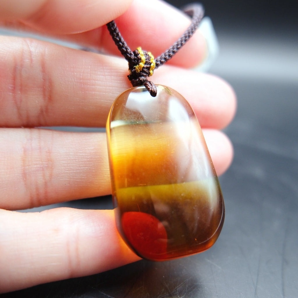 Pendant: transparent Baltic amber, yellow top shading to a dark red base, rounded rectangle, dark cord, held in fingers