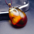 Pendant: transparent Baltic amber teardrop, orange-yellow shading to dark red, dark cord, leaning on a metal tray