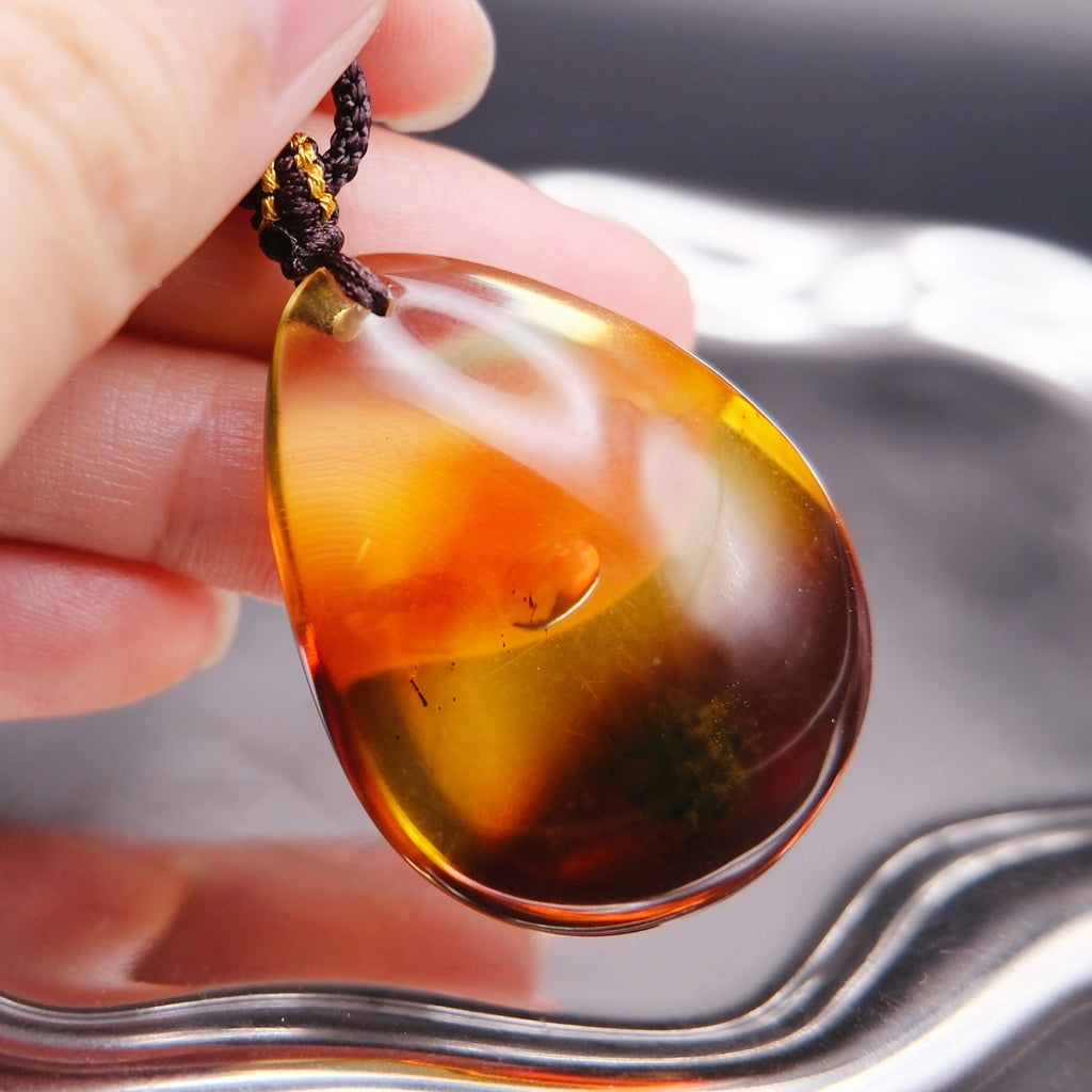 Pendant: transparent Baltic amber teardrop, orange-yellow shading to dark red, dark cord, held in fingers