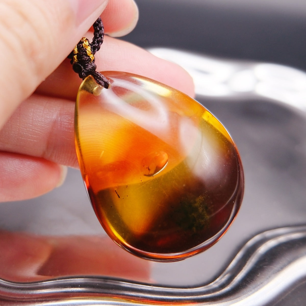 Pendant: transparent Baltic amber teardrop, orange-yellow shading to dark red, dark cord, held in fingers