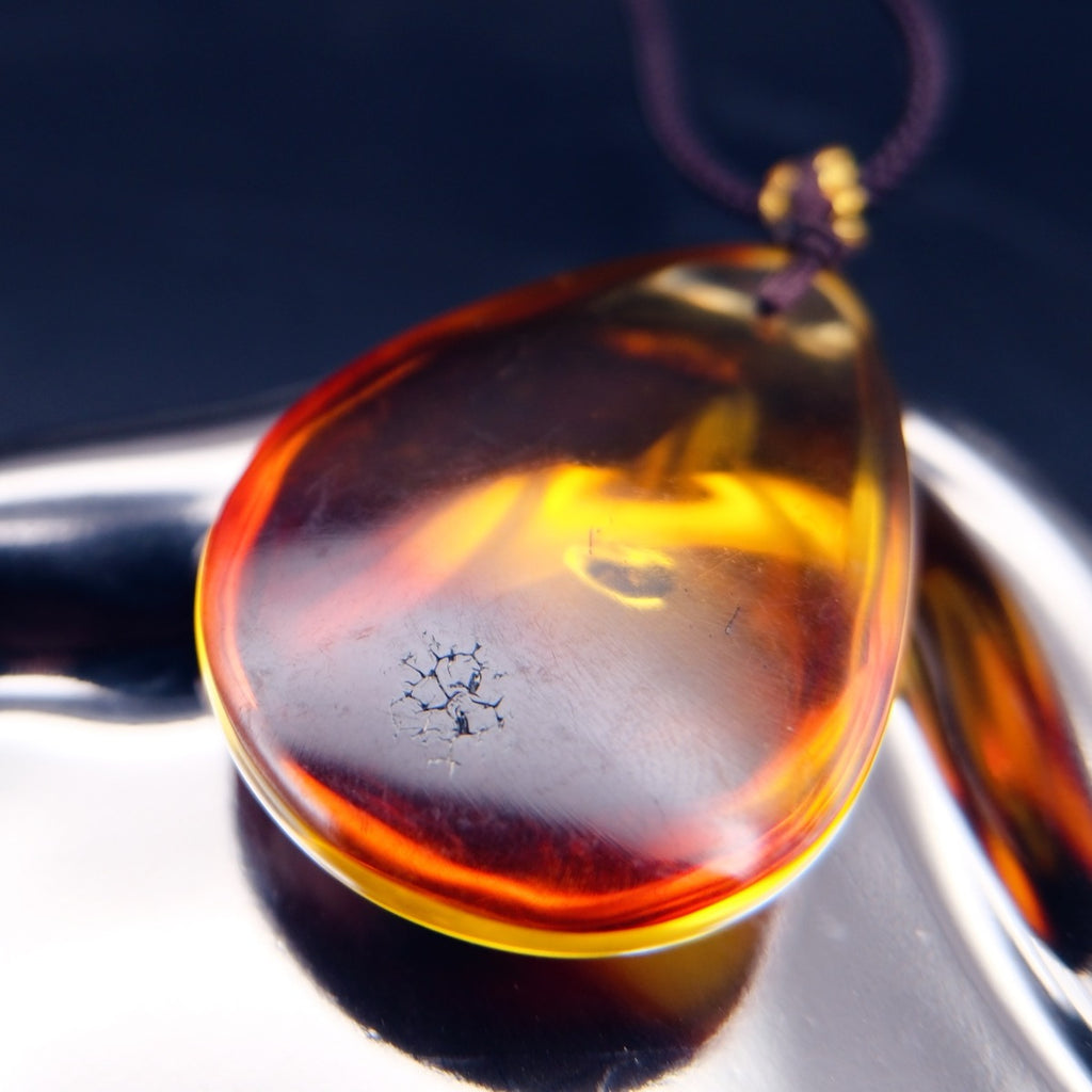 Pendant: transparent Baltic amber teardrop, orange-yellow shading to dark red, dark cord, lying flat