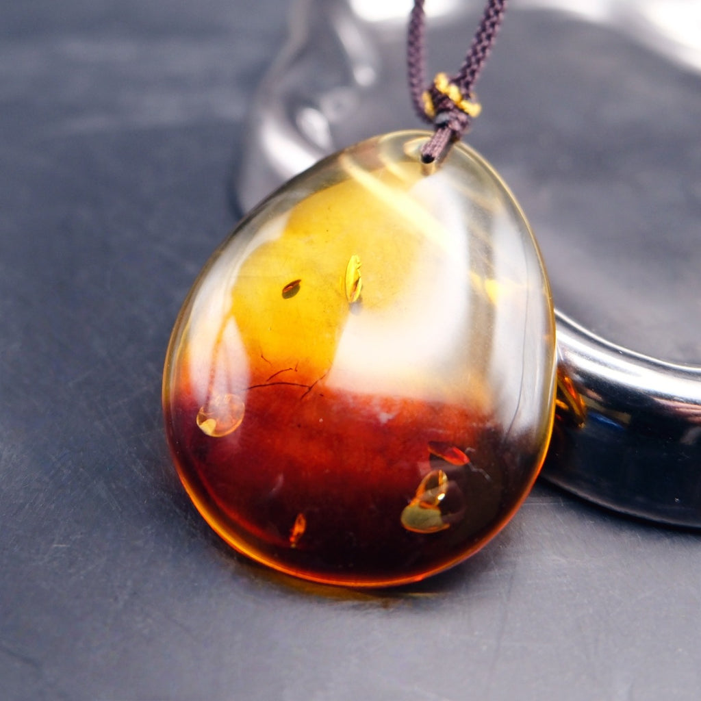 Pendant: transparent Baltic amber teardrop, yellow top and dark red lower half, dark cord, leaning on a metal tray